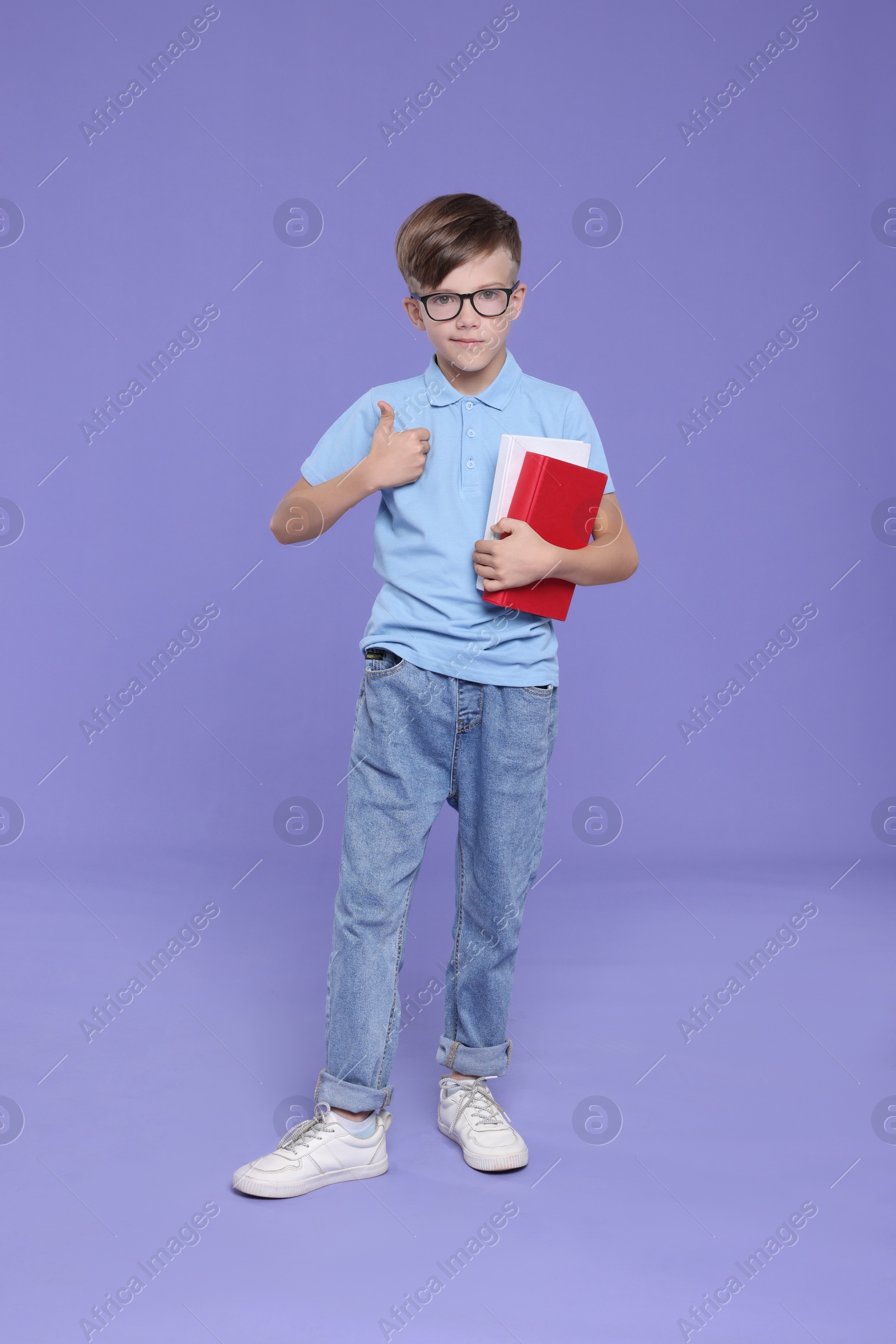 Photo of Cute schoolboy in glasses holding books and showing thumbs up on violet background