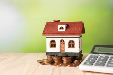 Photo of House model, calculator and coins on wooden table against blurred green background, space for text