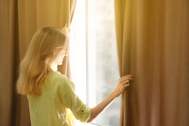 Young woman opening window curtains at home