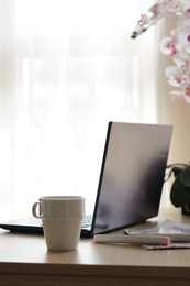 Ceramic cup, laptop and stationery on wooden table indoors. Good morning