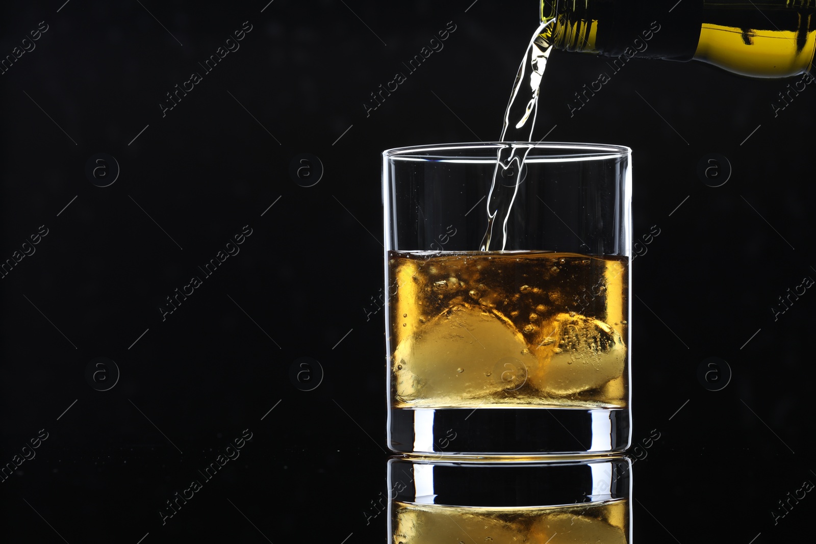 Photo of Pouring tasty whiskey from bottle into glass with ice at mirror table against black background, closeup. Space for text