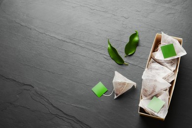 Photo of Box with paper tea bags and leaves on black table, flat lay. Space for text