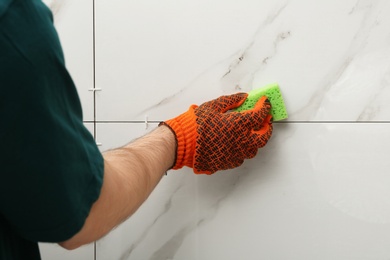 Photo of Man cleaning joint between ceramic tiles with sponge on wall, closeup. Building and renovation works