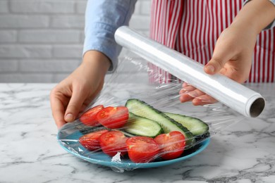 Photo of Woman putting plastic food wrap over plate of fresh vegetables at white marble table indoors, closeup