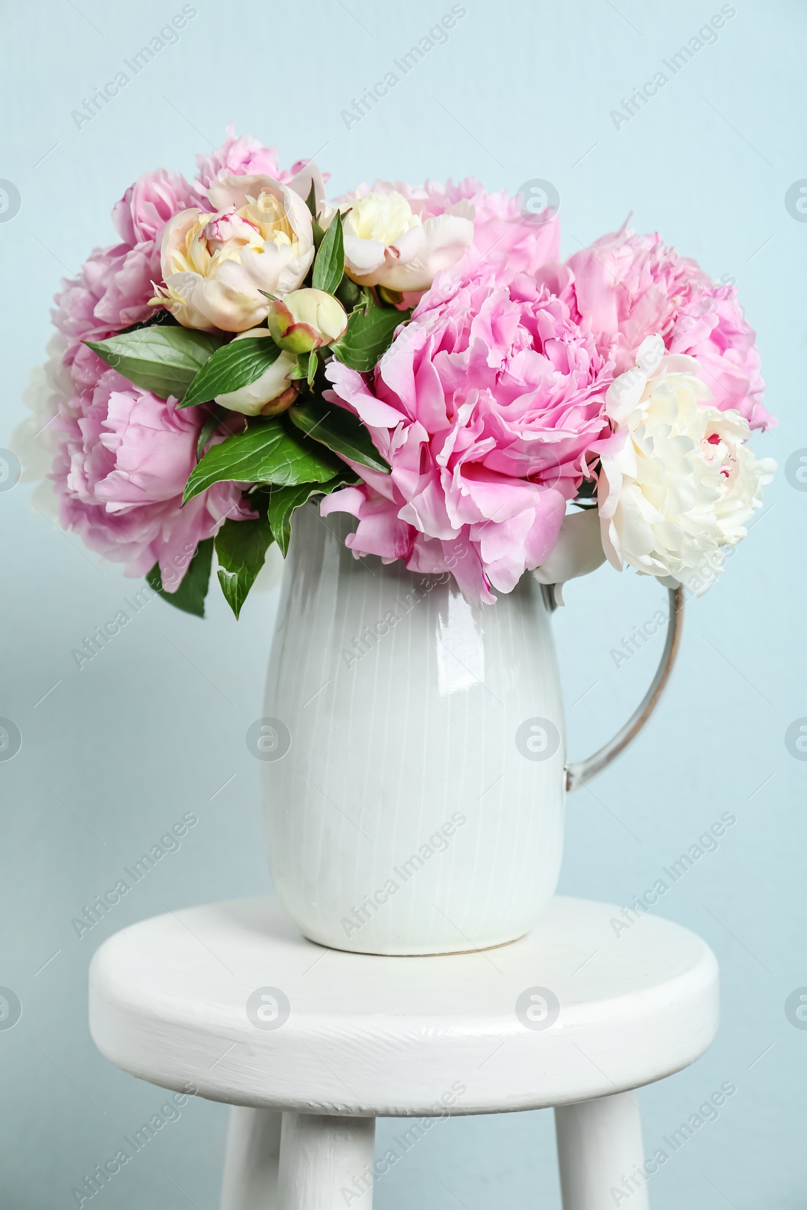 Photo of Beautiful peonies in jug on white stool against light blue background