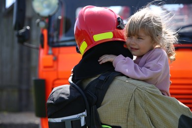 Firefighter in uniform holding rescued little girl near fire truck outdoors. Save life