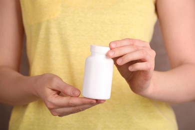 Woman holding blank white bottle with vitamin pills against light brown background, closeup