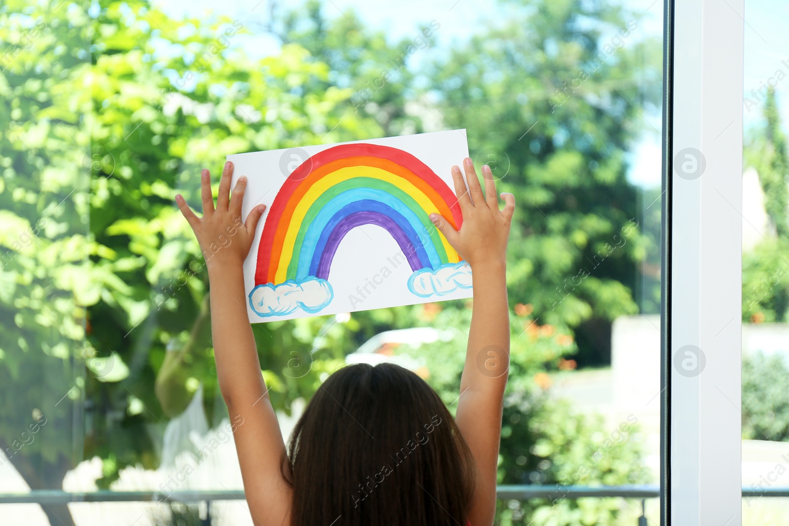 Photo of Little girl with picture of rainbow near window indoors.  Stay at home concept