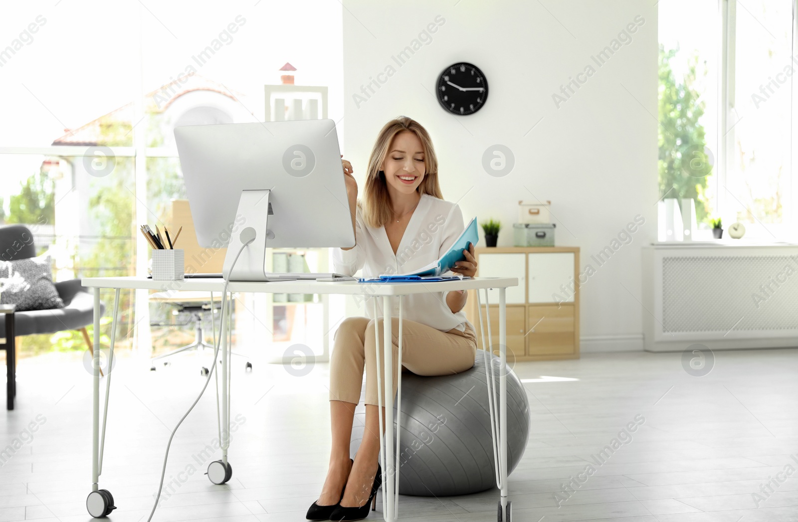 Photo of Happy young businesswoman sitting on fitness ball at desk in office. Workplace exercises