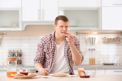 Photo of Young man eating tasty toasted bread with jam at table in kitchen