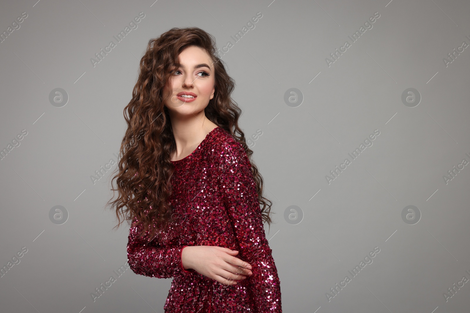 Photo of Beautiful young woman with long curly brown hair in pink sequin dress on grey background, space for text