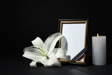 Photo of Funeral photo frame with ribbon, white lily and candle on dark table against black background. Space for design