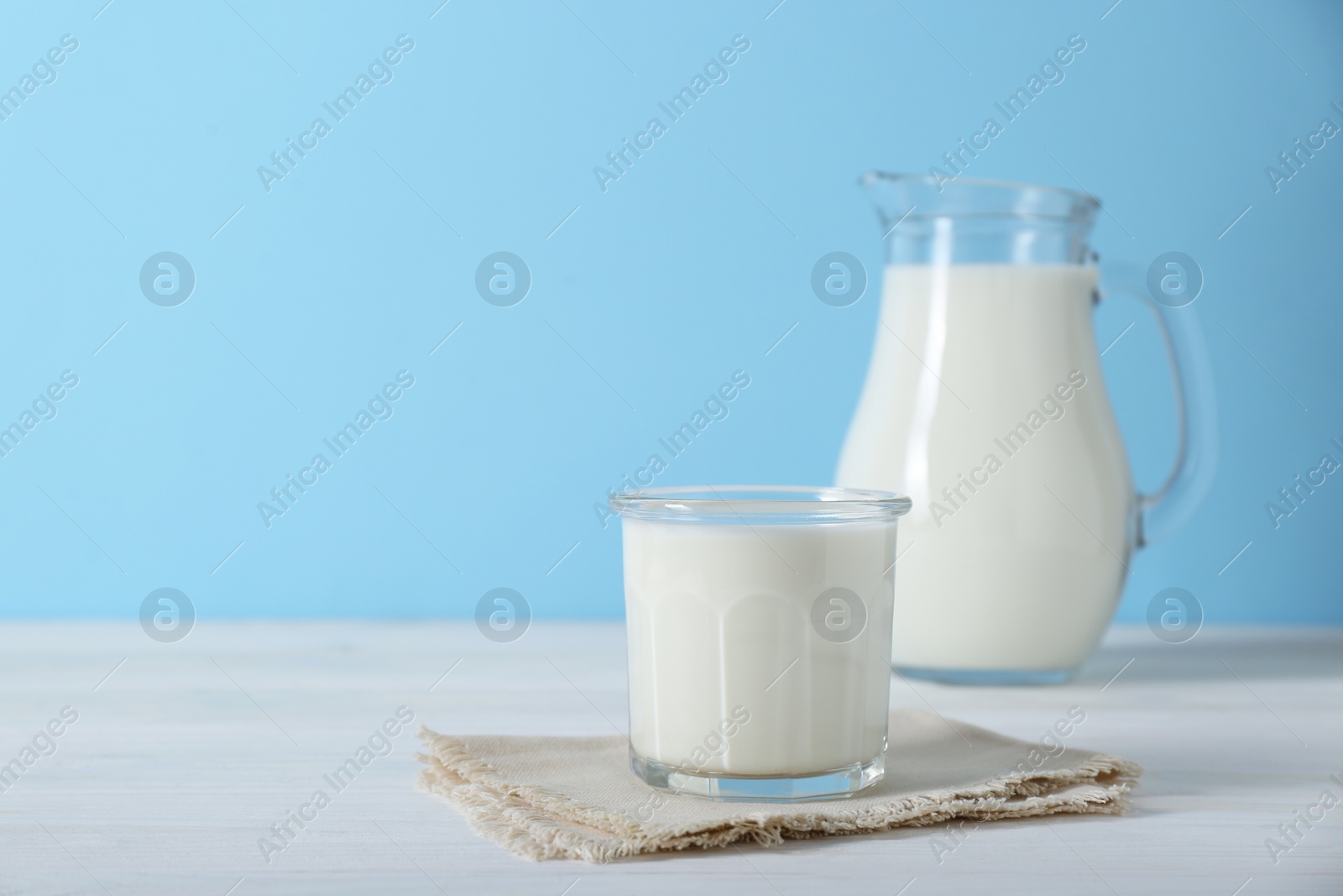 Photo of Glass of fresh milk and jug on white wooden table against light blue background, space for text