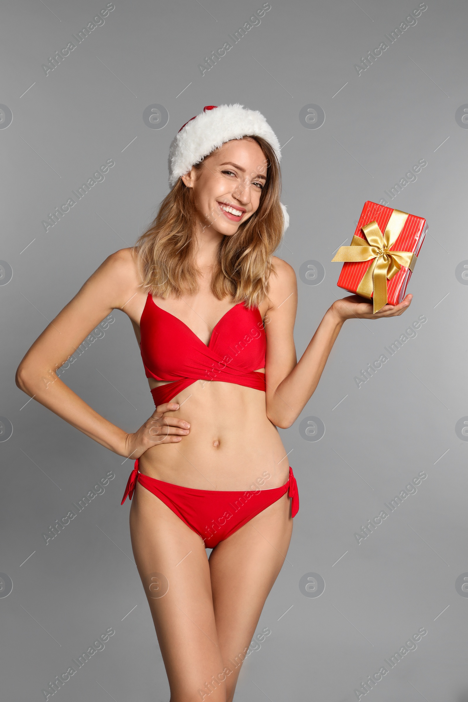 Photo of Happy young woman in red bikini and Santa hat with gift box on light grey background. Christmas celebration