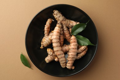 Photo of Bowl with raw turmeric roots and green leaves on light brown table, flat lay