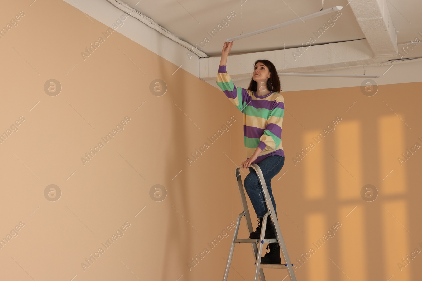 Photo of Young woman installing ceiling lamp on stepladder indoors, space for text