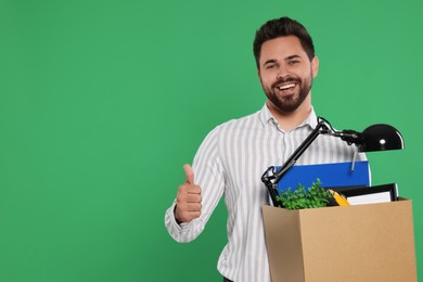 Photo of Happy unemployed man with box of personal office belongings showing thumb up on green background. Space for text