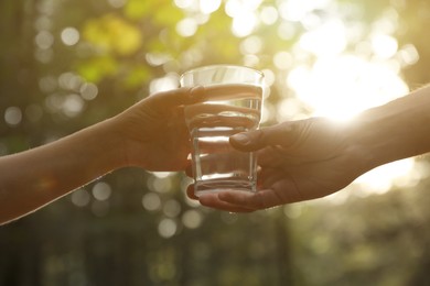 Photo of Man giving woman glassfresh water in forest on sunny day, closeup