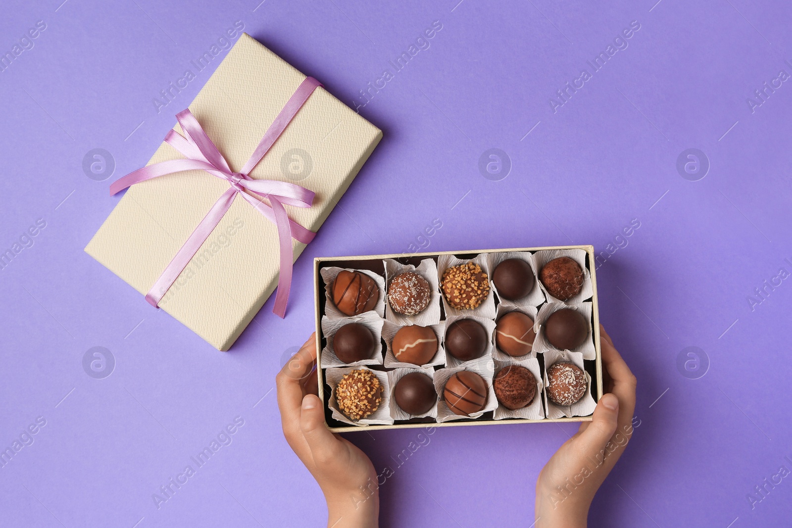 Photo of Child with box of delicious chocolate candies on light purple background, top view