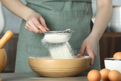 Woman sieving flour into bowl at table in kitchen, closeup