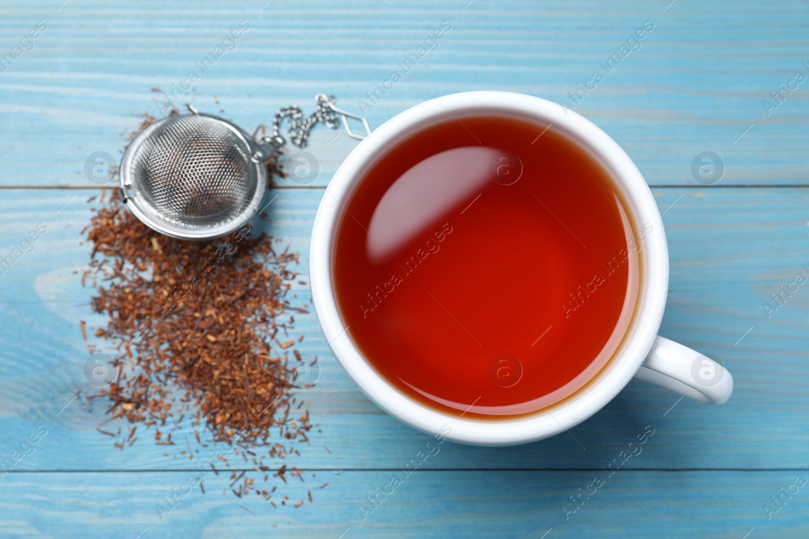 Photo of Ceramic cup of aromatic rooibos tea, infuser and scattered dry leaves on light blue wooden table, flat lay