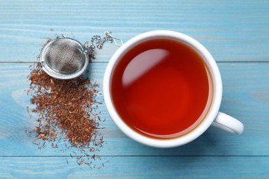 Photo of Ceramic cup of aromatic rooibos tea, infuser and scattered dry leaves on light blue wooden table, flat lay