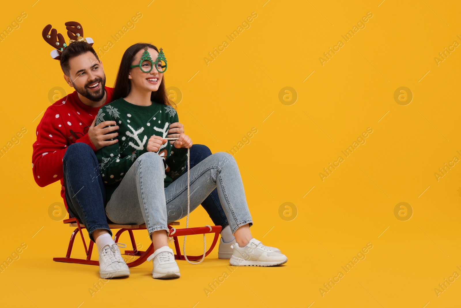 Photo of Happy young couple in Christmas sweaters, reindeer headband and party glasses on sled against orange background. Space for text