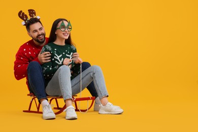 Photo of Happy young couple in Christmas sweaters, reindeer headband and party glasses on sled against orange background. Space for text
