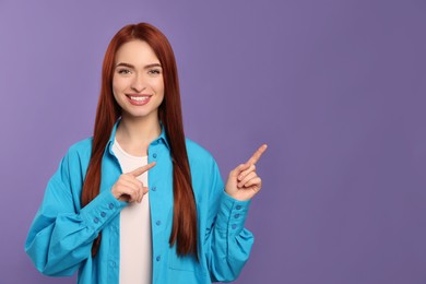 Photo of Happy woman with red dyed hair pointing somewhere on purple background, space for text
