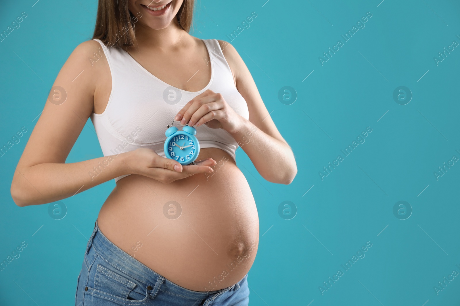Photo of Young pregnant woman holding alarm clock near her belly on light blue background, closeup and space for text. Time to give birth