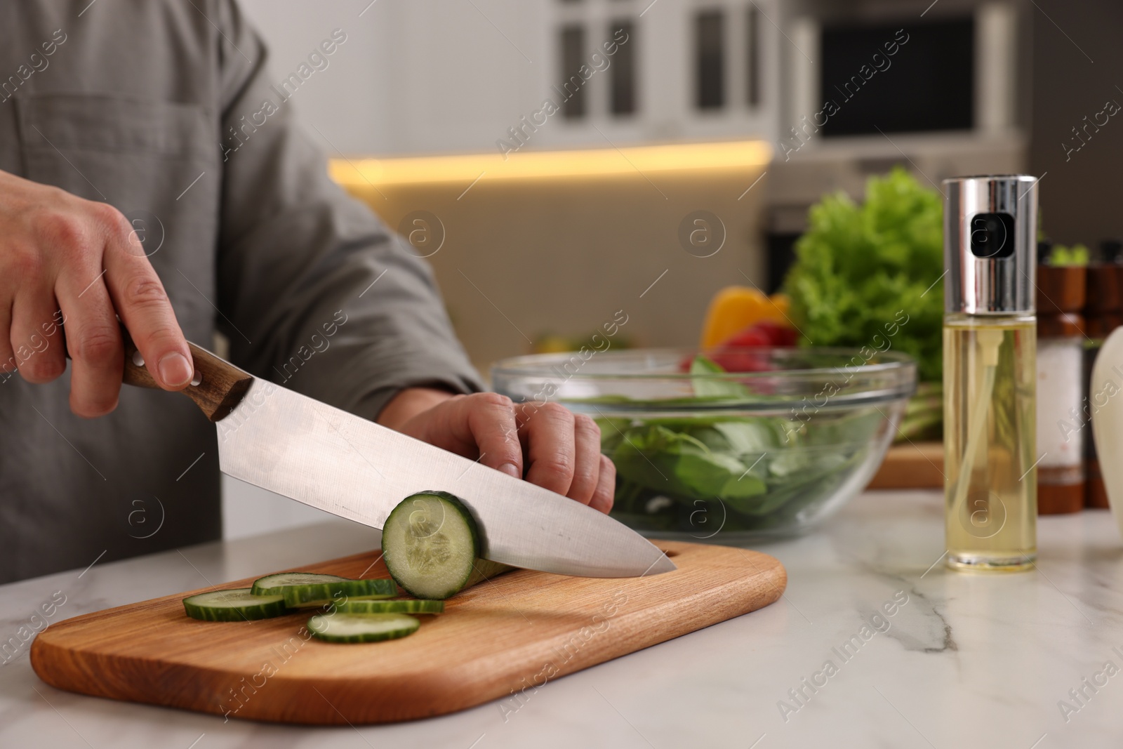 Photo of Cooking process. Man cutting fresh cucumber at white marble countertop in kitchen, closeup