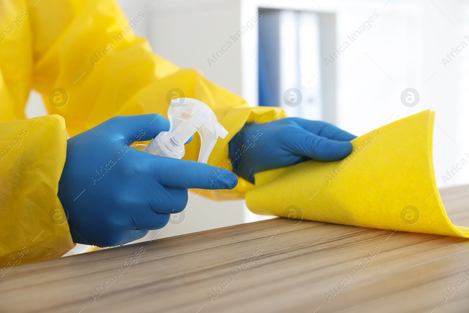 Photo of Employee in protective suit and gloves sanitizing wooden countertop indoors, closeup. Medical disinfection