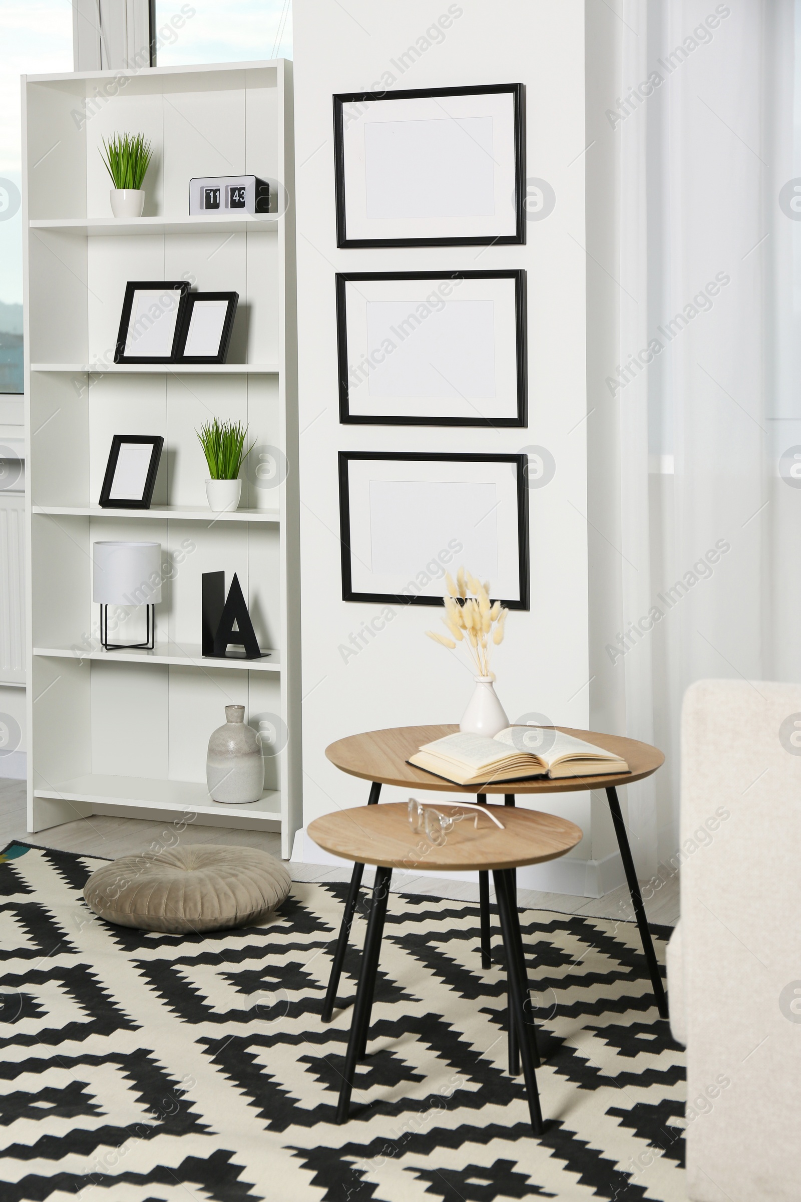 Photo of Stylish room interior with empty frames hanging on white wall near shelving unit