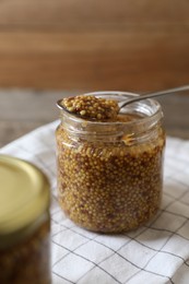 Jar and spoon of whole grain mustard on white checkered tablecloth, closeup