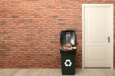 Photo of Full trash bin near brick wall indoors, space for text. Waste recycling