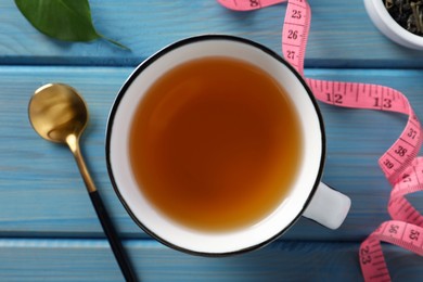 Cup of herbal diet tea and measuring tape on light blue wooden table, flat lay. Weight loss concept