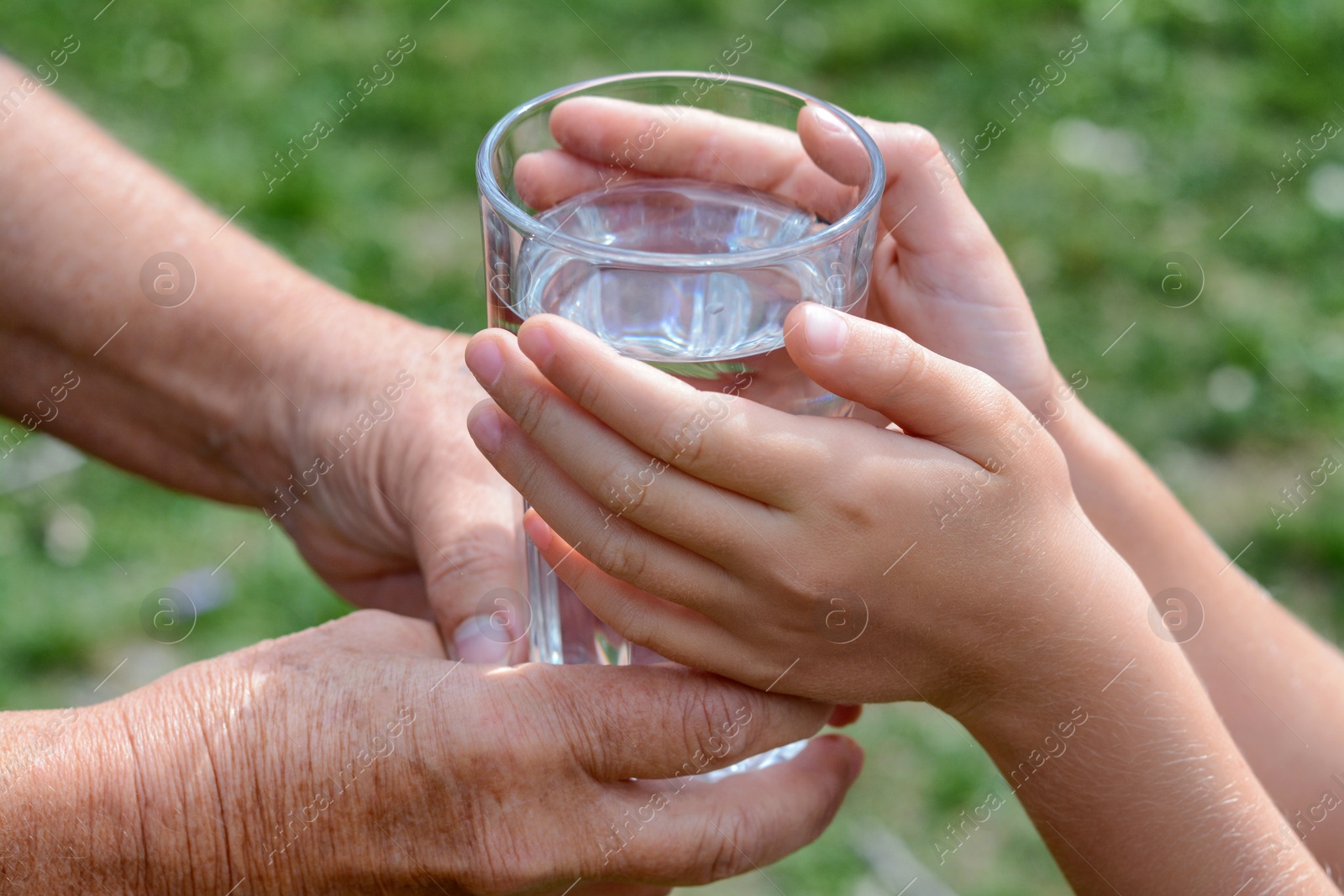 Photo of Child giving glass of water to elderly woman outdoors, closeup