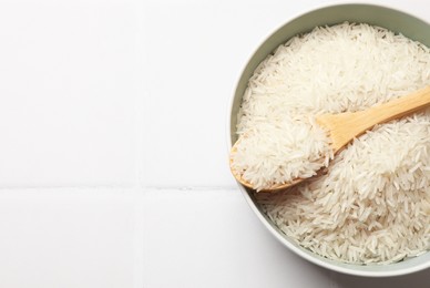 Photo of Raw basmati rice in bowl and spoon on white tiled table, top view. Space for text