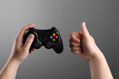 Woman using wireless game controller and showing thumbs up on grey background, closeup