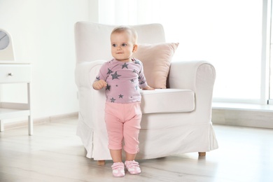 Cute baby girl standing near armchair in room