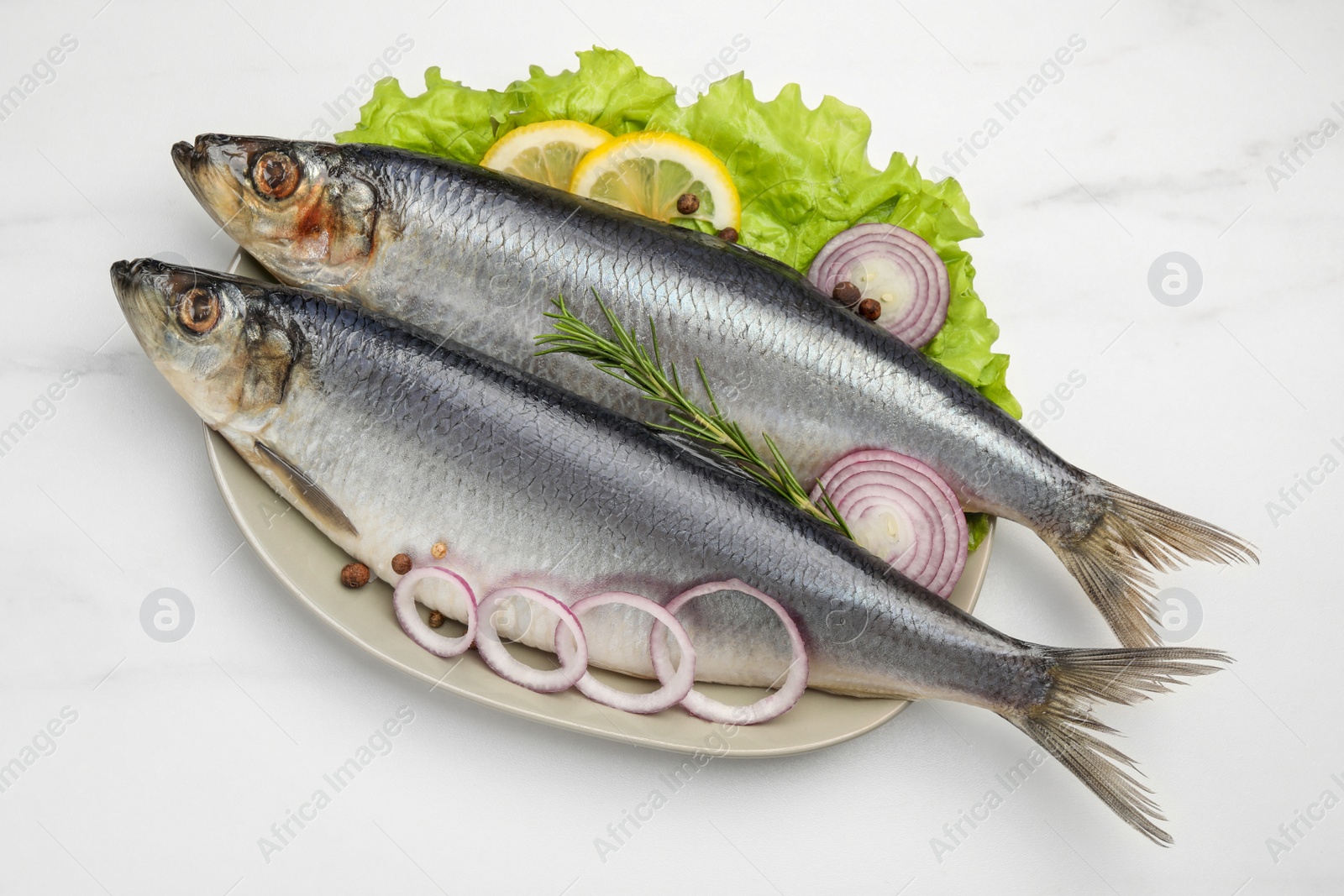 Photo of Plate with salted herrings, onion rings, slices of lemon, peppercorns and lettuce on white marble table, top view