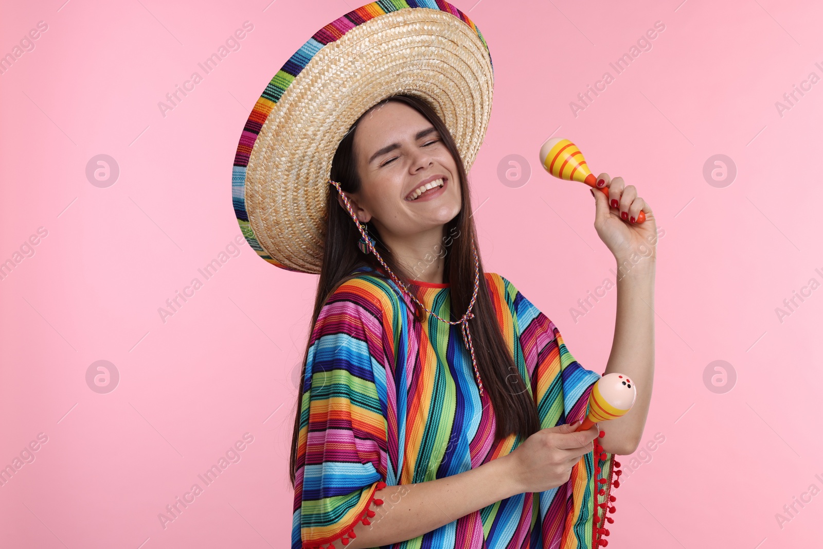 Photo of Young woman in Mexican sombrero hat and poncho dancing with maracas on pink background