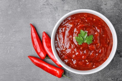 Photo of Bowl of hot chili sauce with parsley and red peppers on grey background, flat lay. Space for text
