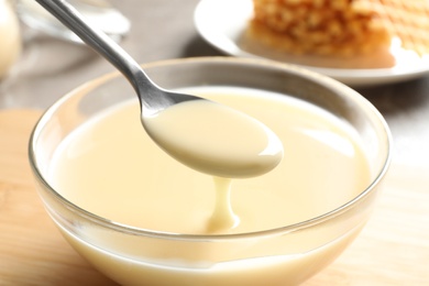 Photo of Spoon of pouring condensed milk over bowl on table, closeup. Dairy products