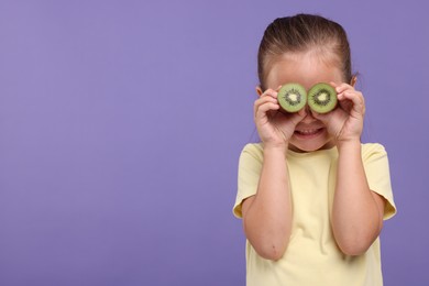 Photo of Smiling girl covering eyes with halves of fresh kiwi on violet background, space for text