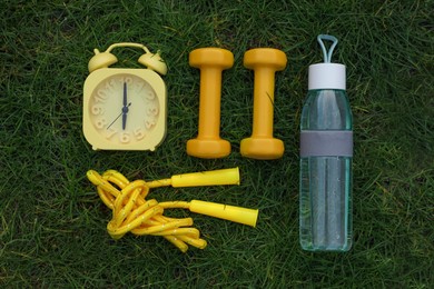 Alarm clock, skipping rope, dumbbells and bottle of water on green grass, flat lay. Morning exercise