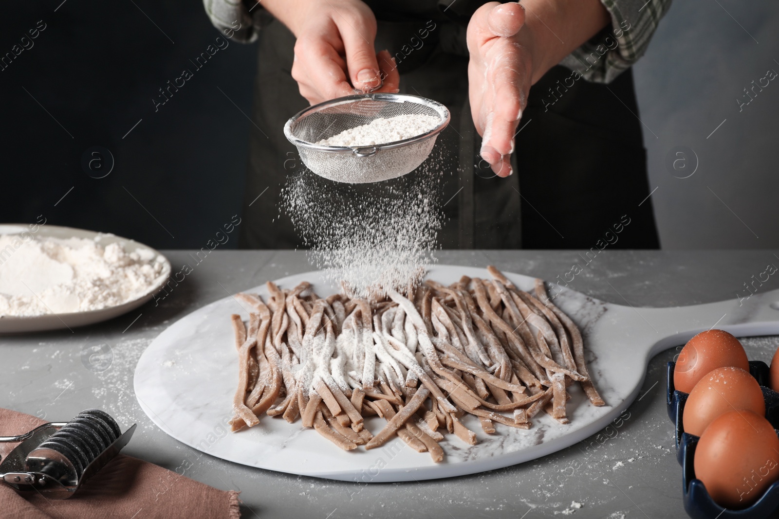 Photo of Woman sprinkling raw soba (buckwheat noodles) with flour at grey table, closeup