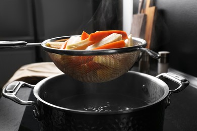 Sieve with cut parsnips and carrots over pot of boiling water in kitchen, closeup