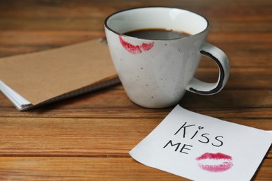 Notebook, sticky note with phrase Kiss Me, lipstick mark and cup of coffee on wooden table