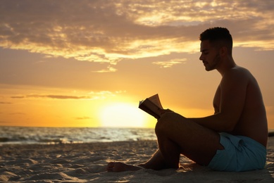 Young man reading book on sandy beach near sea